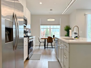 Una cocina moderna con armarios blancos, electrodomésticos de acero inoxidable, un comedor con mesa y sillas, y un ventanal que ofrece vistas al exterior.