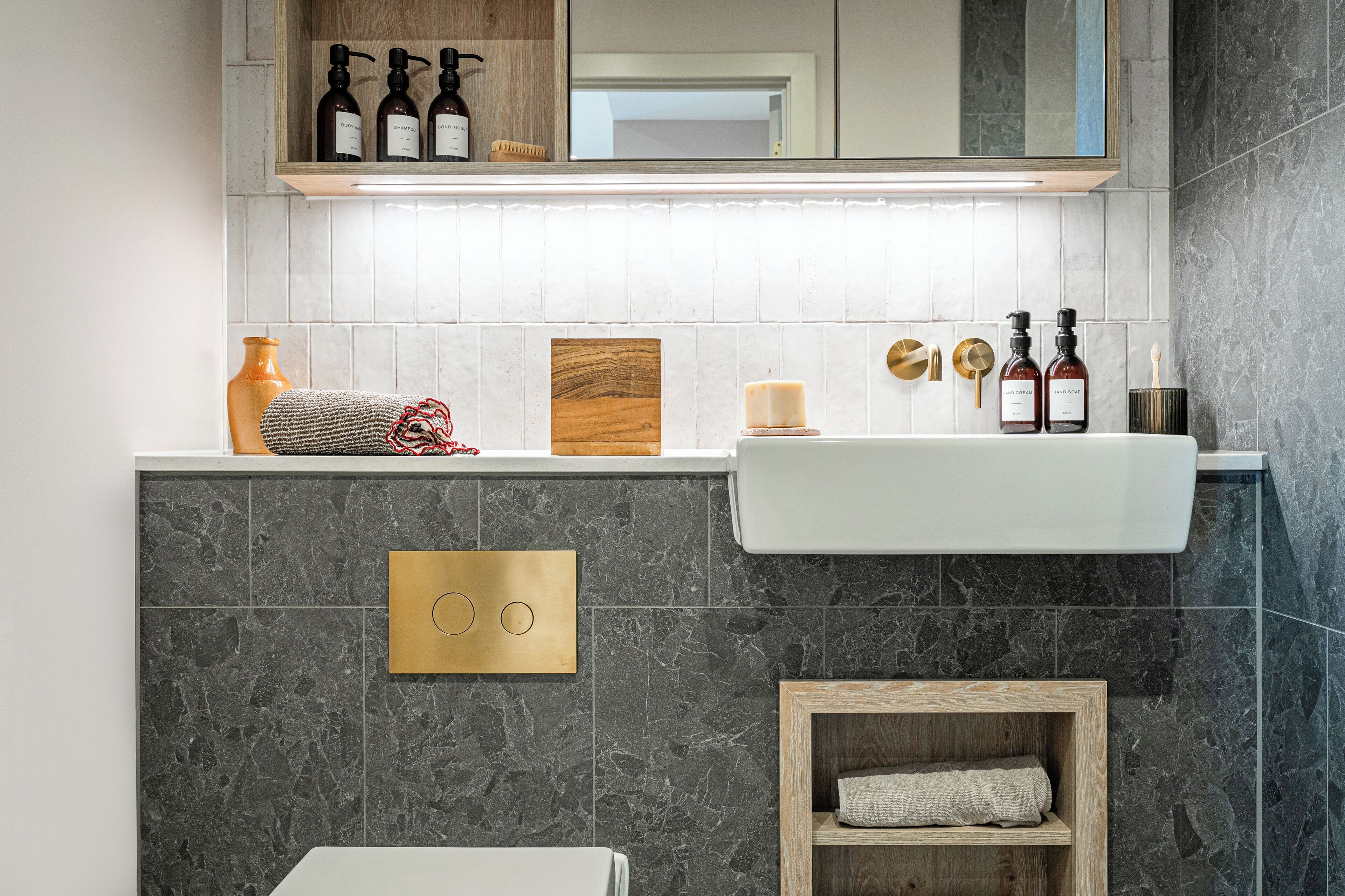 A modern bathroom with a white sink, dark marble tiles, a wooden shelf with toiletries, and a mirror above the sink.