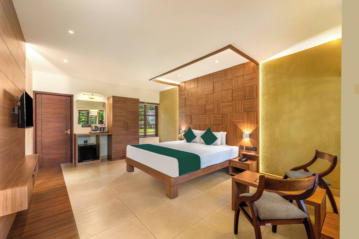 A hotel room with a wooden bed, a desk with a chair, a TV, and a bathroom visible through a glass door.