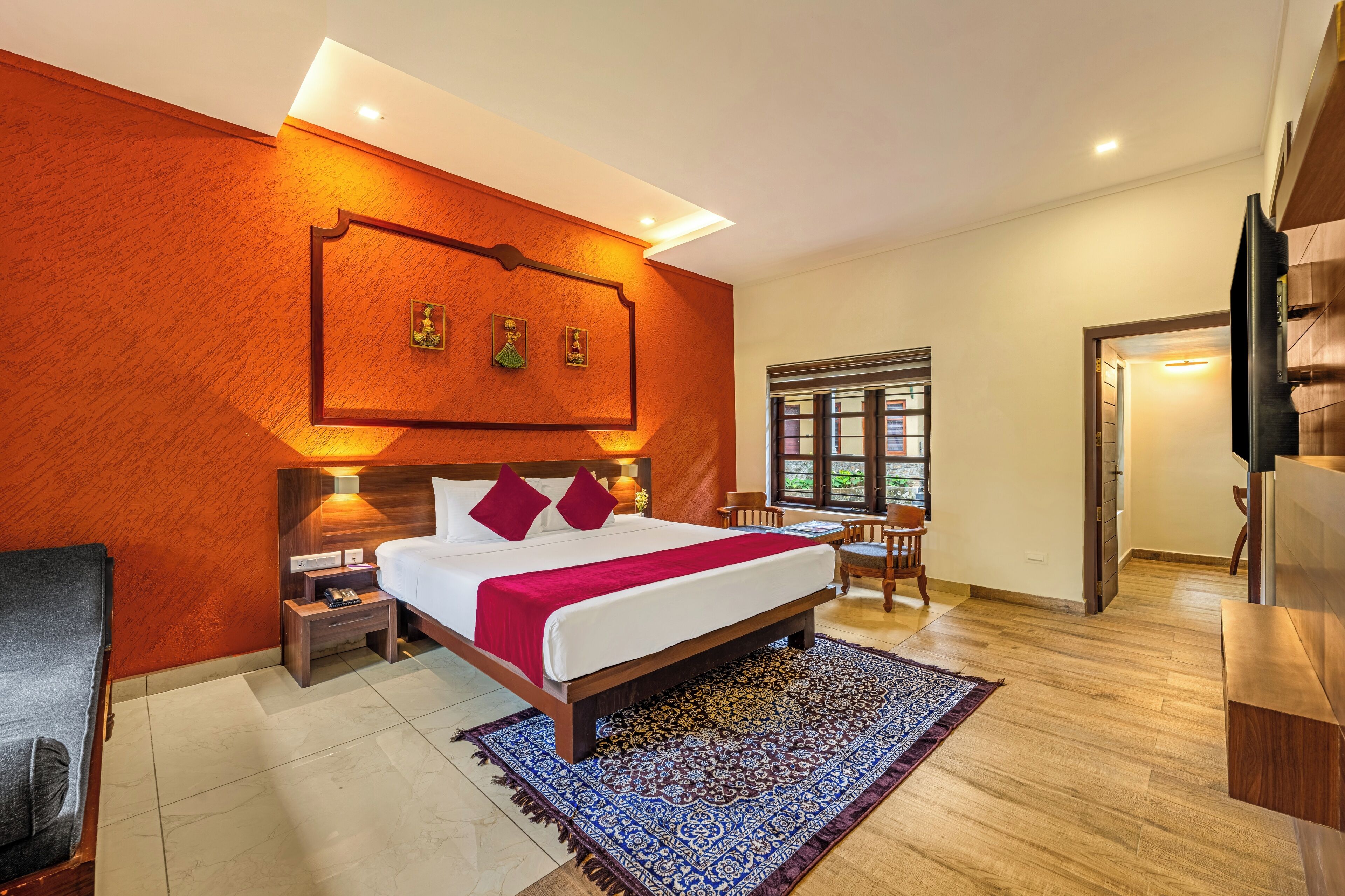 A modern hotel room with a large bed, wooden flooring, a red accent wall, a small desk, and a television.