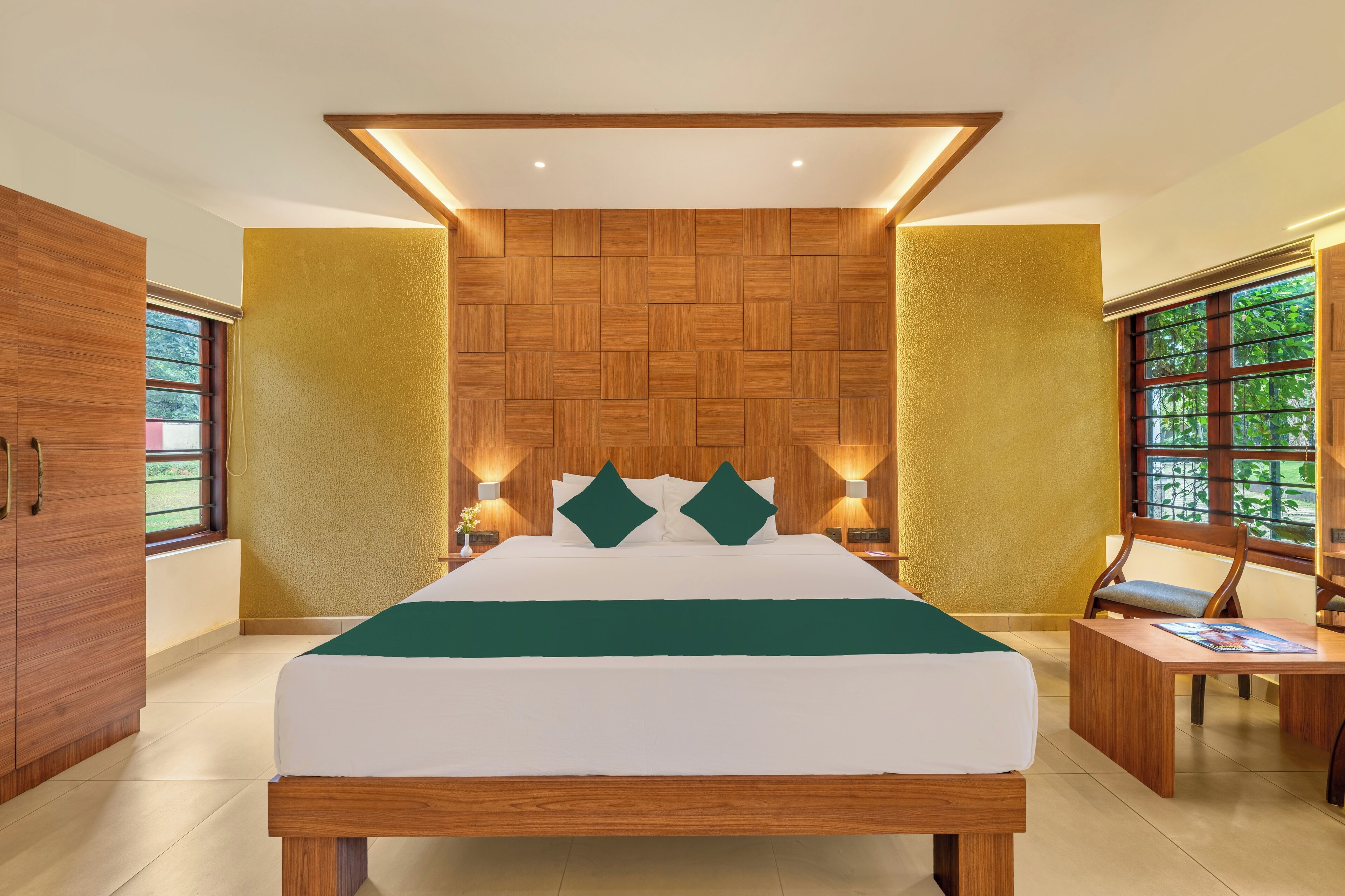 A modern bedroom with a wooden headboard, a large bed with a green bedspread, bedside tables with lamps, and a view of greenery through the windows.