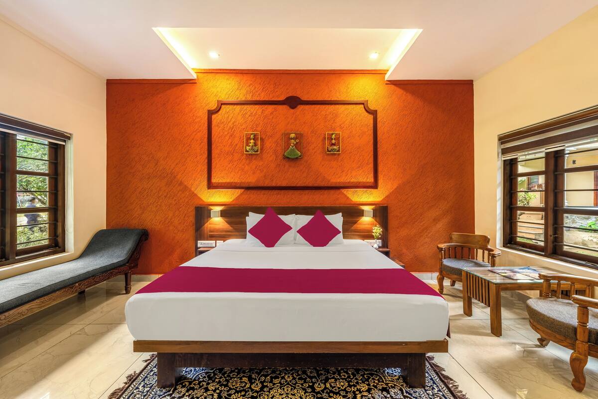 A hotel room with a large bed, wooden headboard, and a red accent wall. There are two windows with wooden frames, a bench, and a table with chairs.