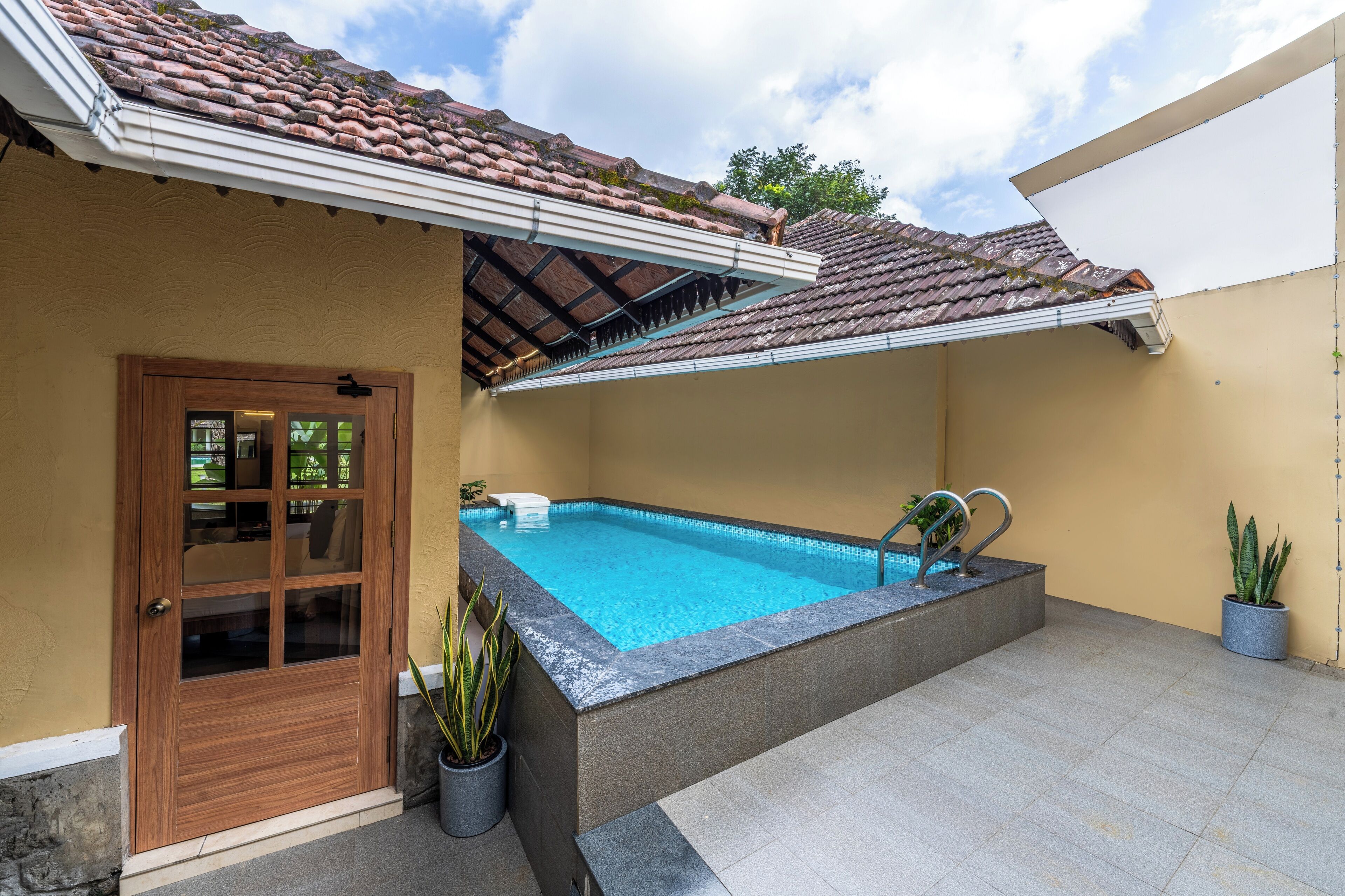 Una piscina con un borde de baldosas, una puerta de madera y plantas en macetas.