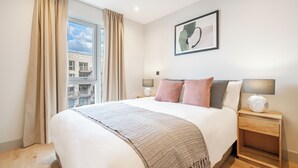 A bedroom with a large bed, two bedside tables with lamps, a window with curtains, and a framed picture on the wall. - Stylish One-bedroom Haven (London)