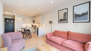 A modern living room with a pink sofa, a purple armchair, and a wooden coffee table. There is a kitchen area with white cabinets and a dining table with chairs. Two framed artworks are displayed on the wall. - Stylish One-bedroom Haven (London)