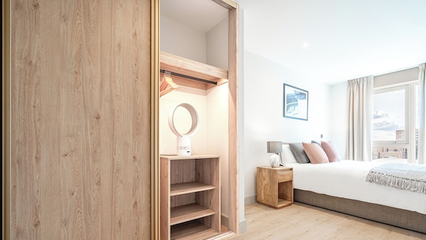 Un dormitorio moderno con un armario de madera, una mesita de noche con lámpara, una cama con muchos cojines y un ventanal con vistas a la ciudad.