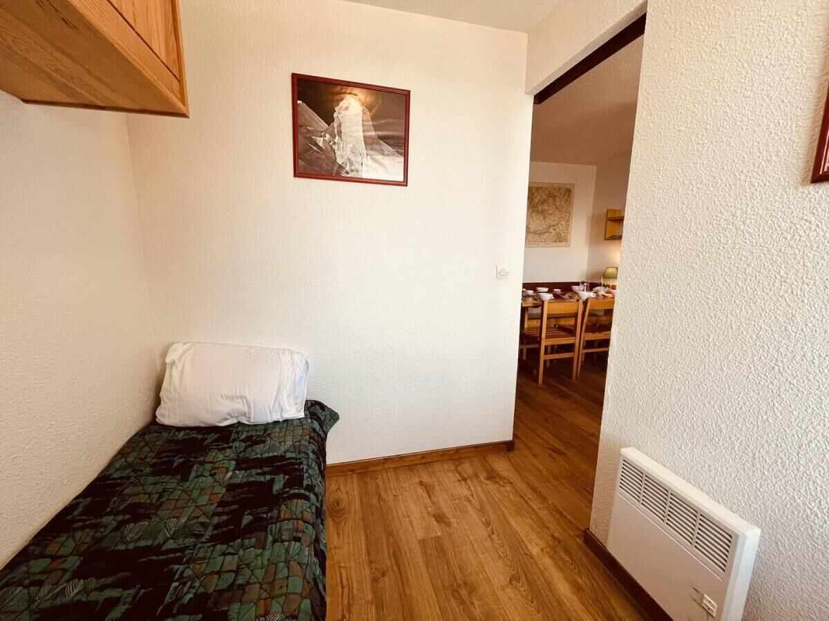 A small, single-bed room with a wooden floor, a white pillow, a framed picture on the wall, and a dining area in the background.