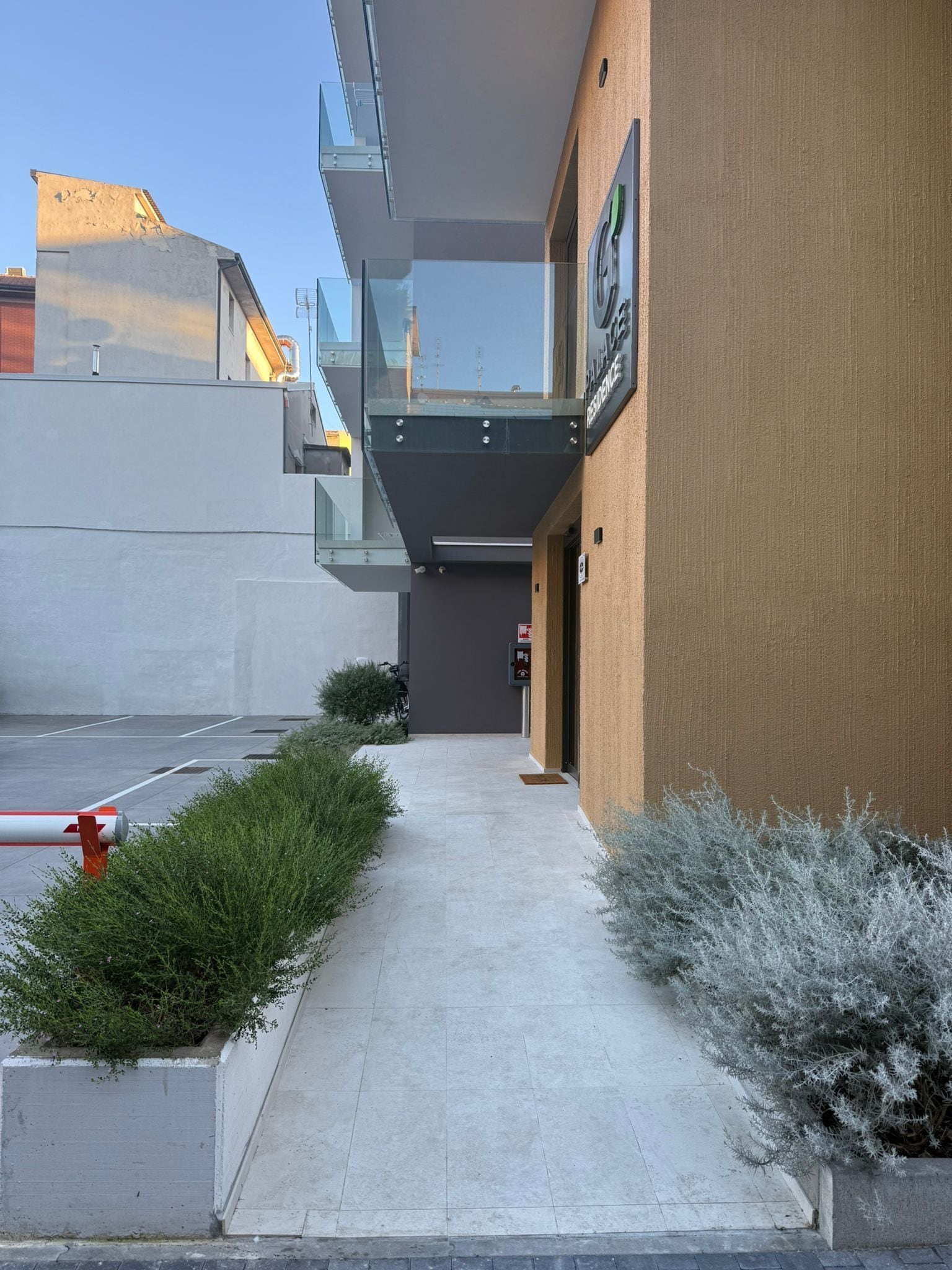 A modern building with a glass balcony, a concrete pathway, and some greenery.