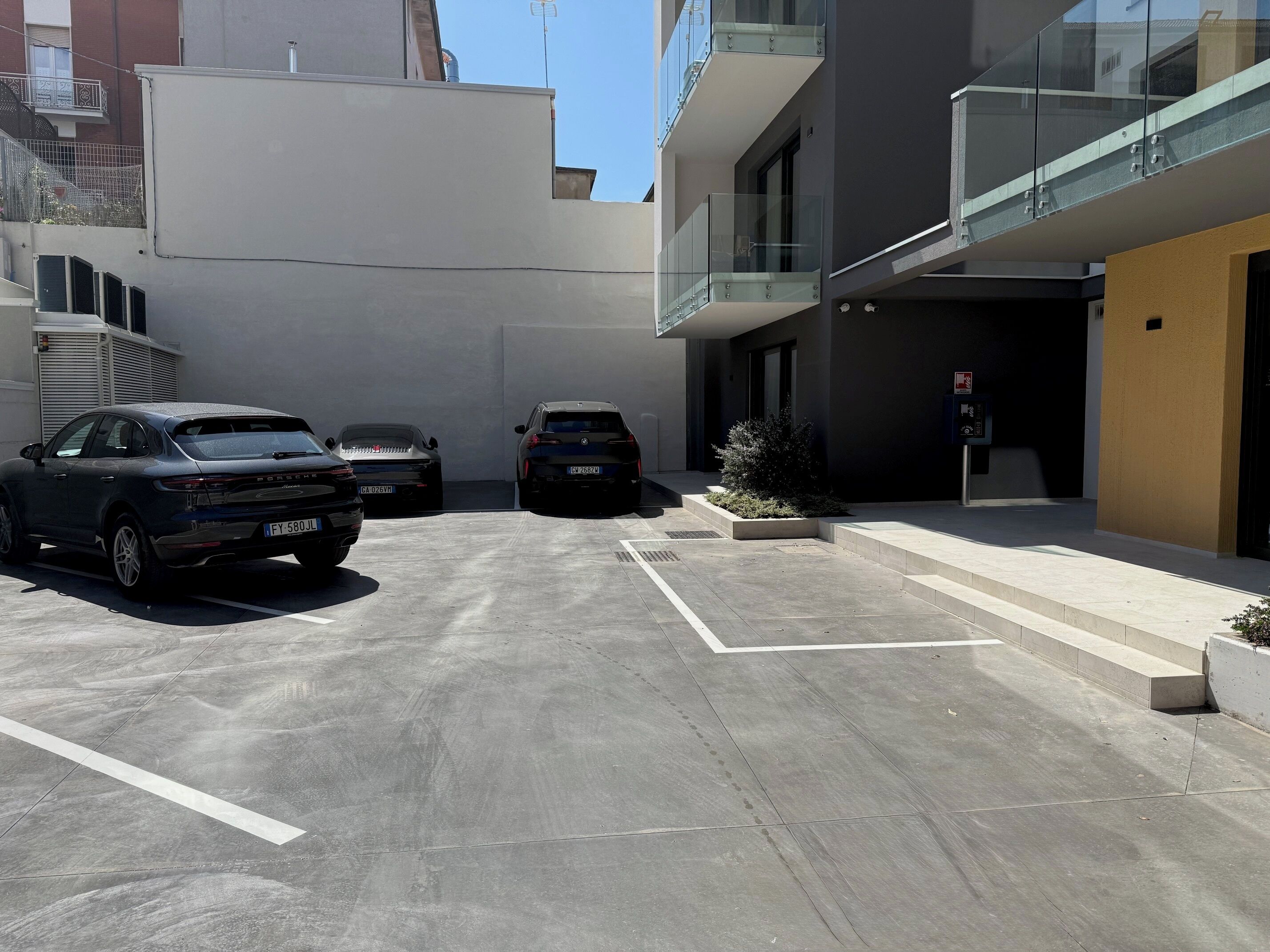 Ein Parkplatz mit zwei Autos, ein Gebäude mit einem Glasbalkon und einer kleinen Pflanze.