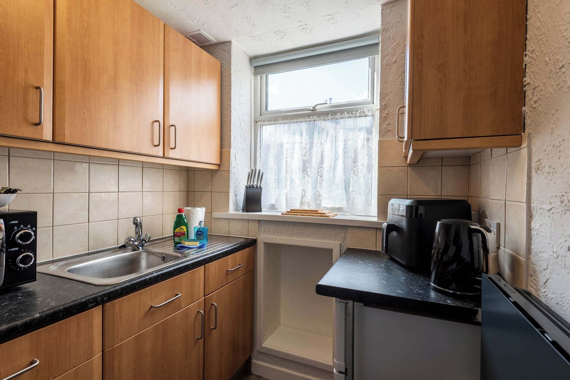 A compact kitchen with wooden cabinets, a sink, a coffee maker, and a window with curtains.
