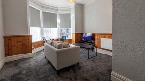 Ein Wohnzimmer mit grauem Sofa, Holzvertäfelung, Fernseher und einem großen Fenster.