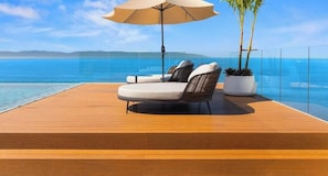 Una terraza de madera con dos sillones reclinables y una sombrilla grande, con vista a un mar azul y cristalino.
