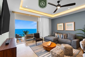 A modern living room with a sofa, coffee table, and chairs, a ceiling fan, and a large window with a view of the ocean.