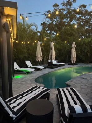 Un área junto a la piscina con sillones de descanso y sombrillas, luces colgantes y una piscina.
