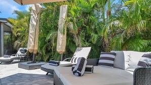 A sunlit outdoor lounge area with wicker furniture, including a sofa and lounge chairs, surrounded by lush tropical foliage. - Paradise Found Home With Private Heated Saltwater Pool And Cabana Plus Game Room (Seminole)
