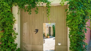 Een houten poort met een silhouet van een vogel, leidend naar een grindpad en een gebouw met een stenen muur en een deur.