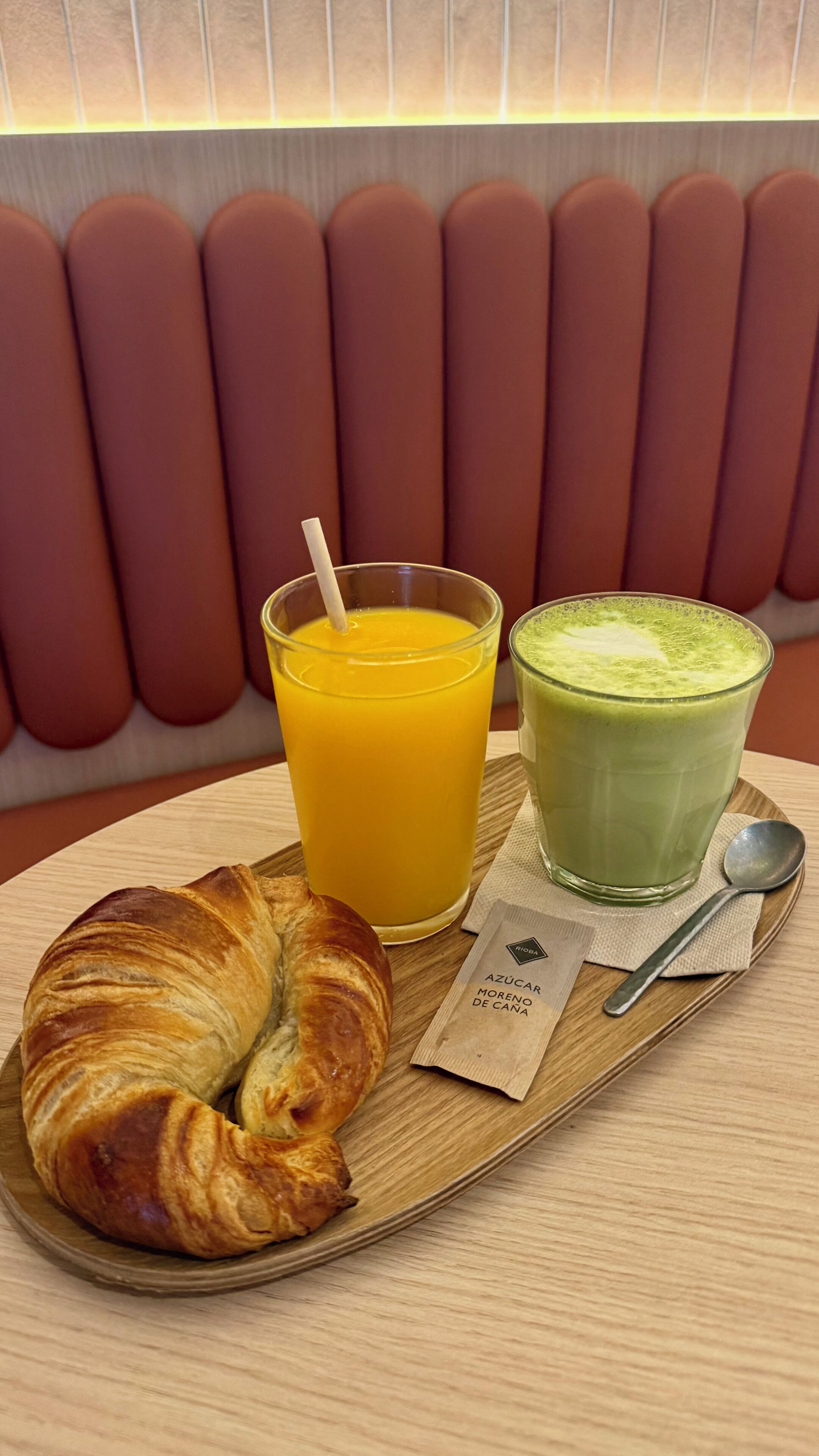 Bandeja de desayuno con un croissant, un vaso de jugo de naranja y un batido verde, sobre una mesa de madera con servilleta y cuchara.