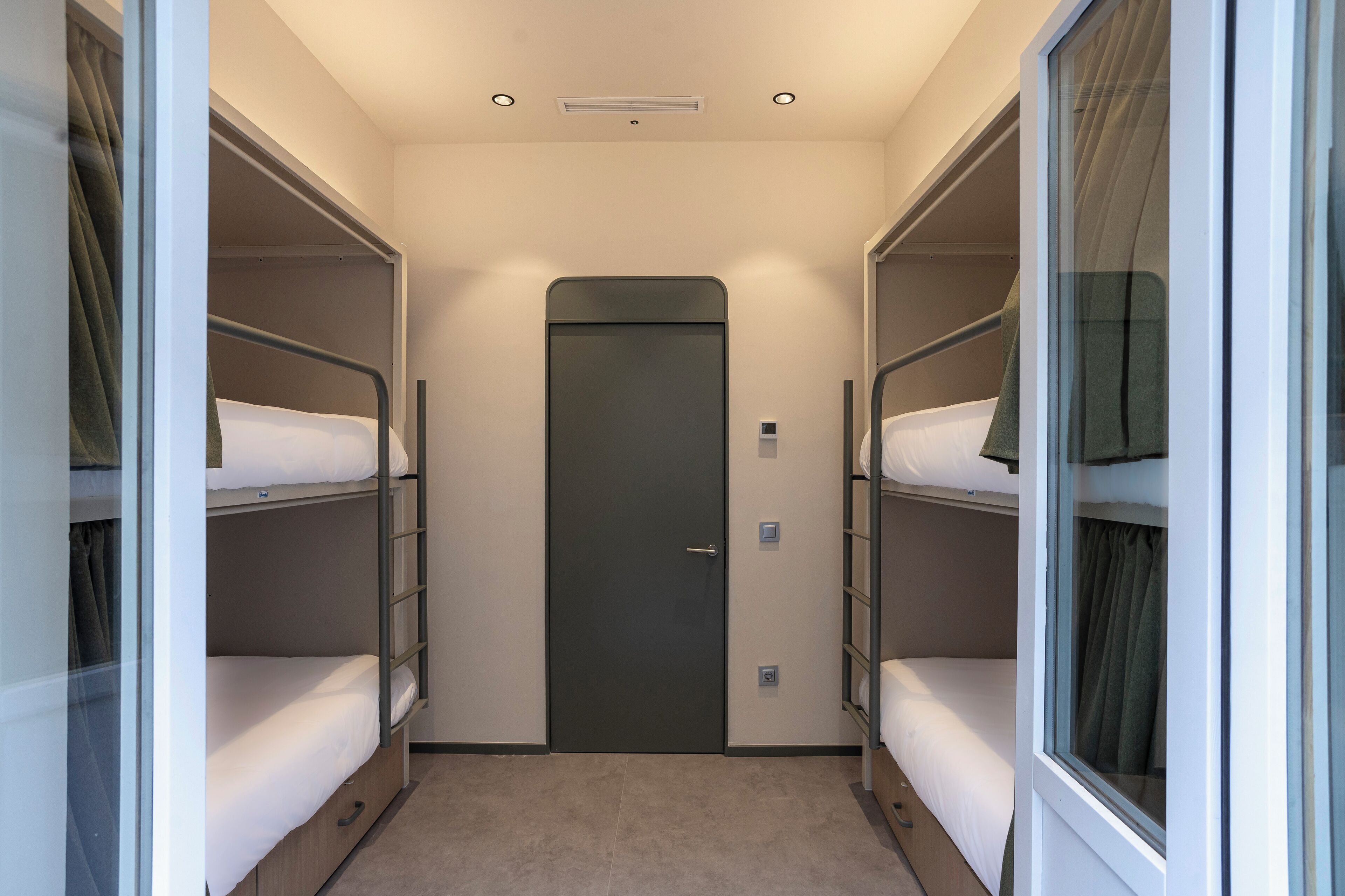 A narrow hallway with bunk beds on either side, a door at the end, and a small window on the right wall.