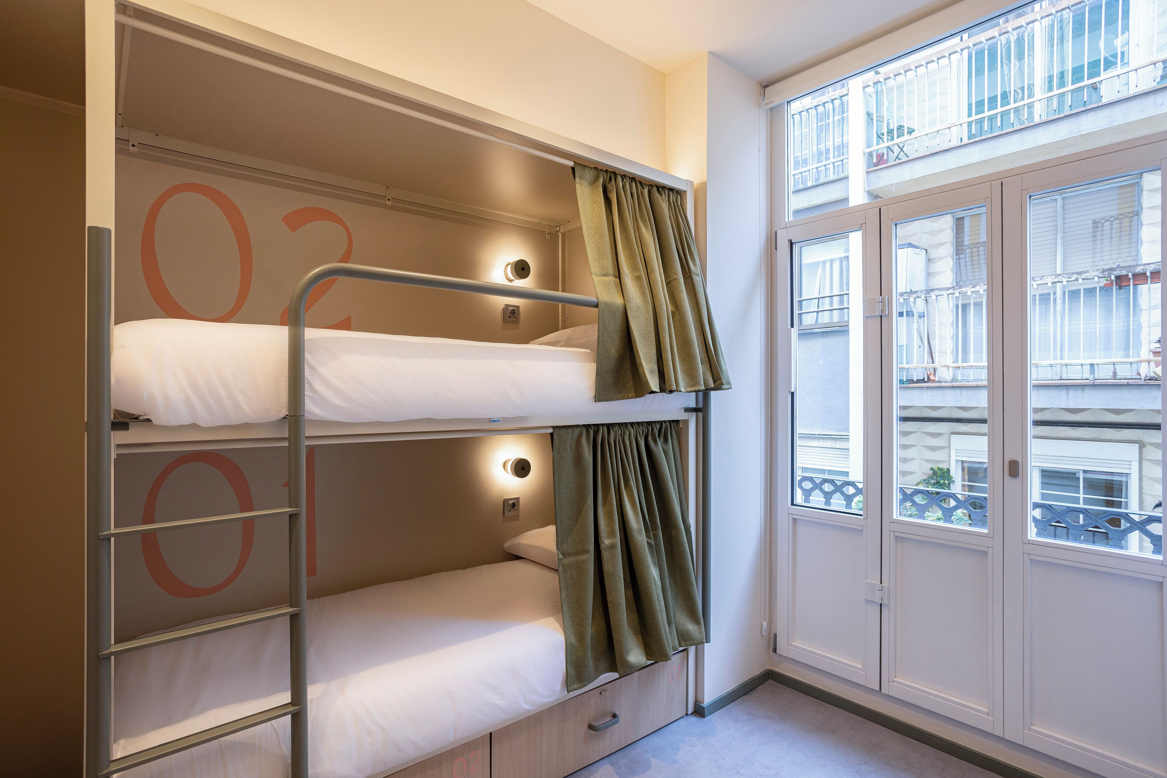 Uma cama beliche com três níveis, uma janela com vista para varandas e uma porta à direita.
