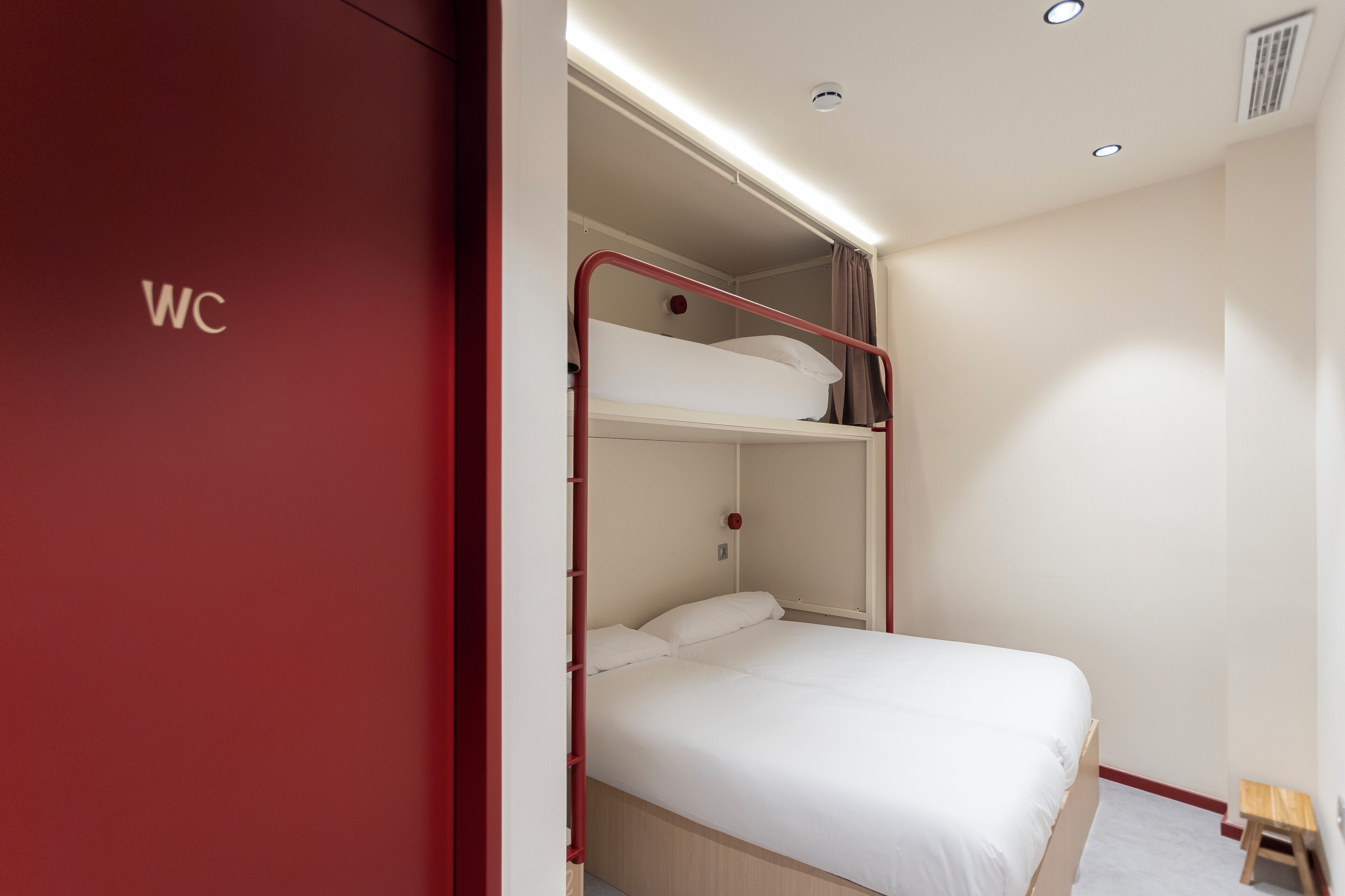 Dormitorio con literas, un armario rojo etiquetado como 'WC' y un taburete de madera pequeño.