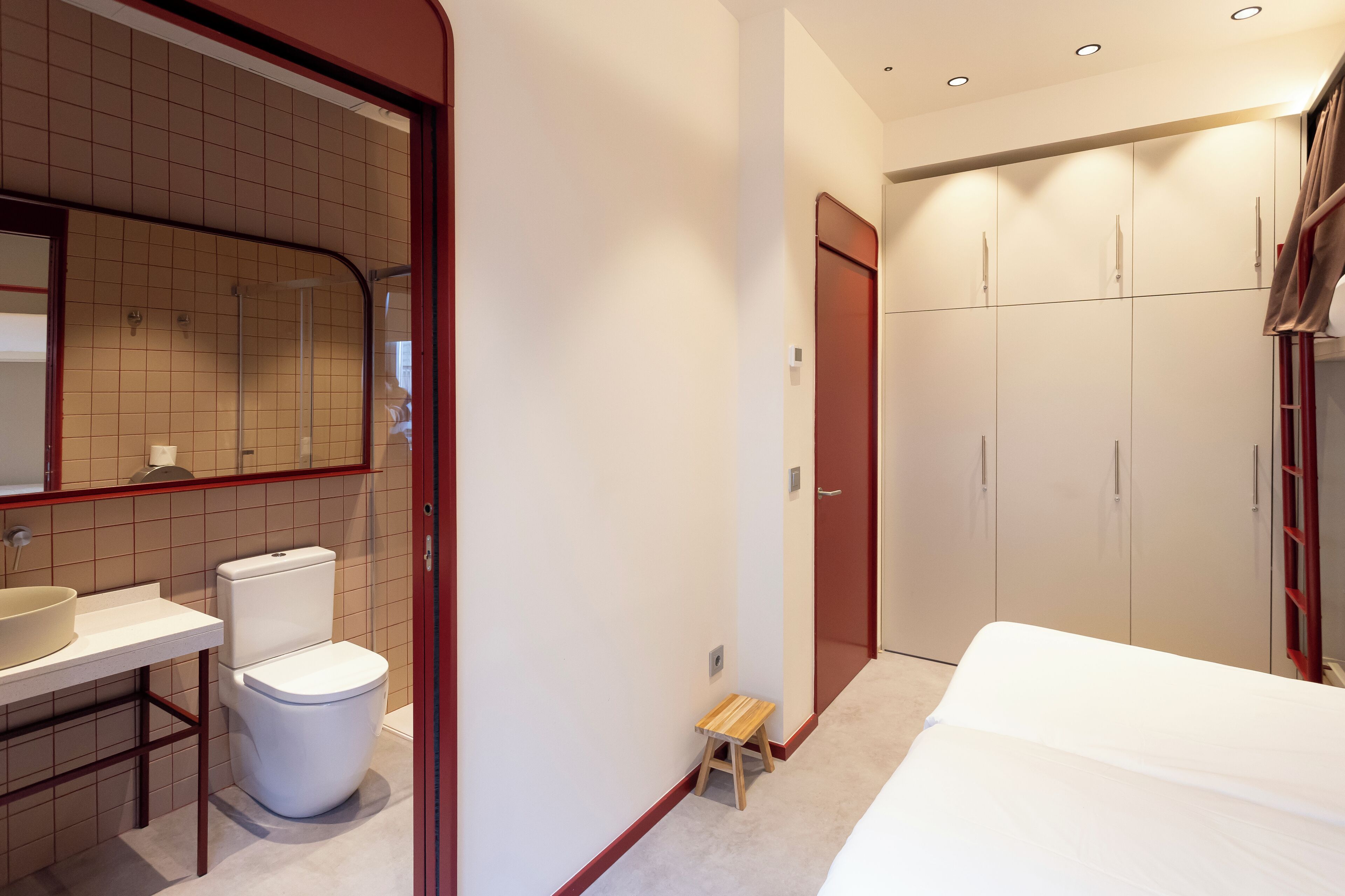 A modern bathroom with a toilet, sink, and mirror. There is a small wooden stool and a bed with a white mattress.