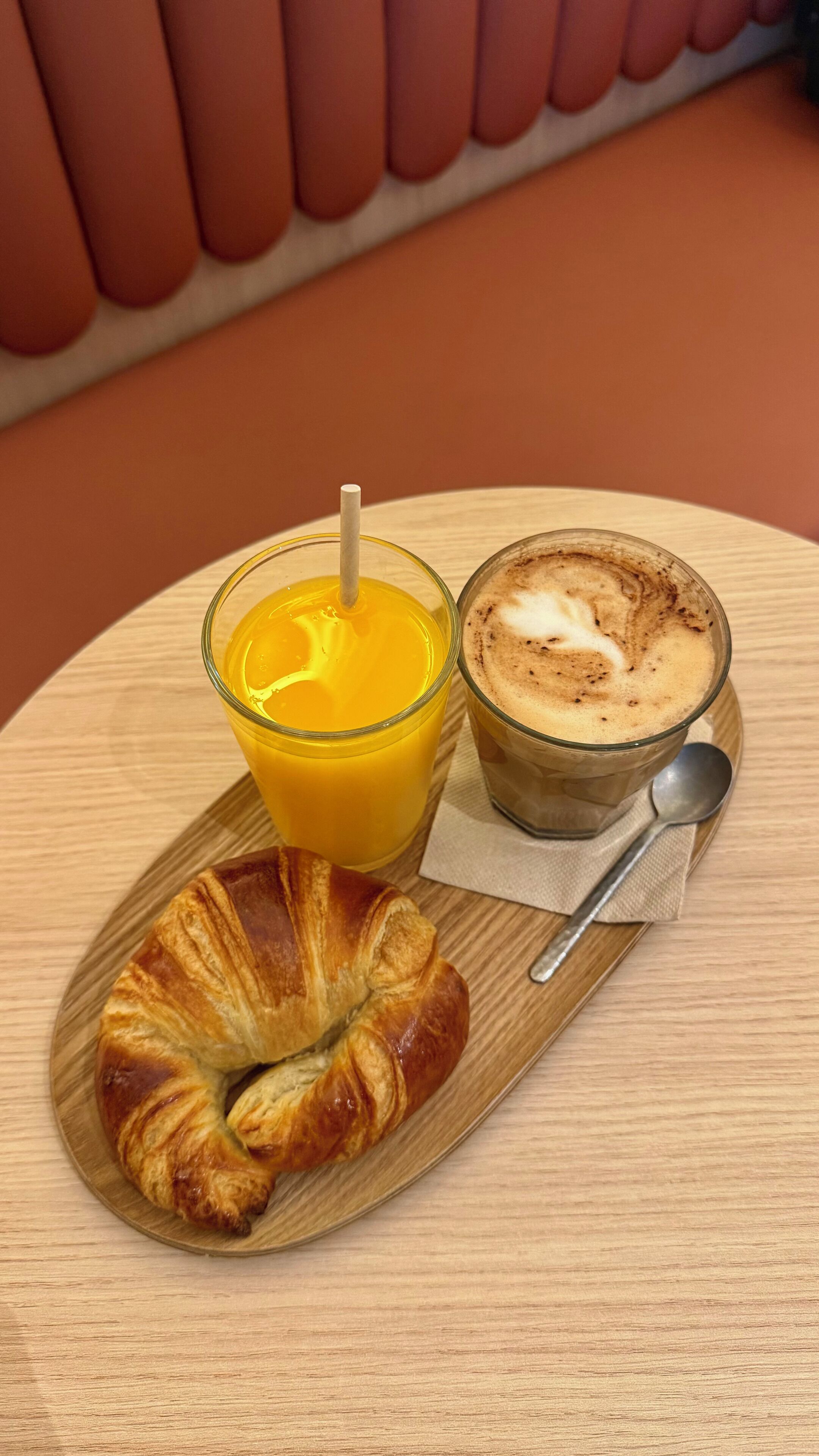 Un croissant sur une planche en bois, un verre de jus d’orange et un verre de café avec une cuillère, le tout posé sur une table ronde.