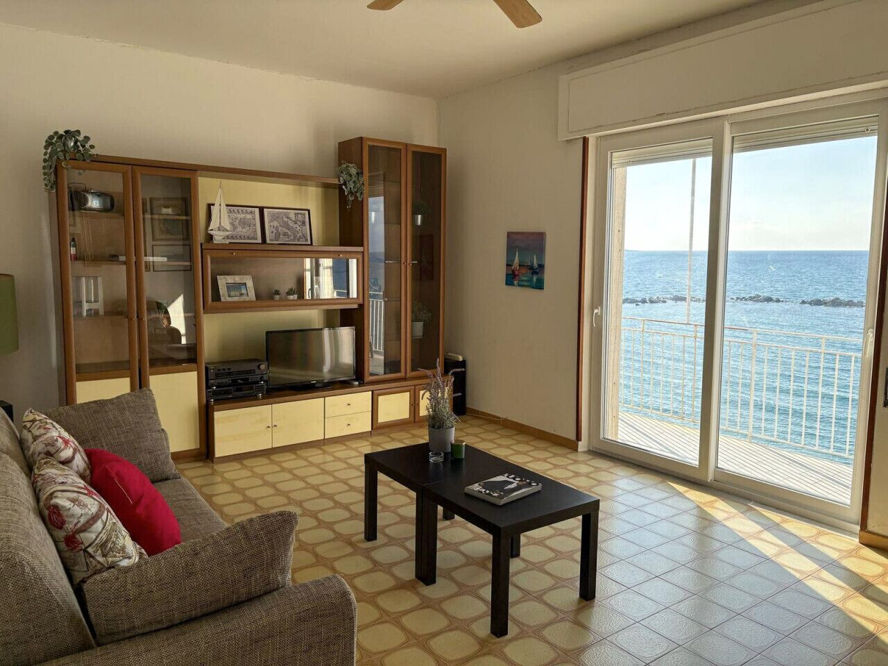 Una sala de estar con un sofá, una mesa de centro, un armario de madera y un ventanal con vista al mar.