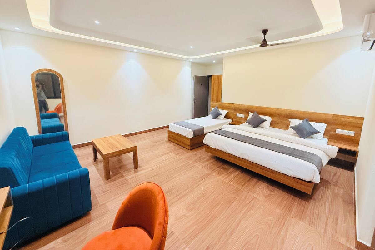 Una habitación de hotel moderna con dos camas individuales, un sofá azul, un sillón naranja, una mesa de centro de madera y un ventilador de techo.