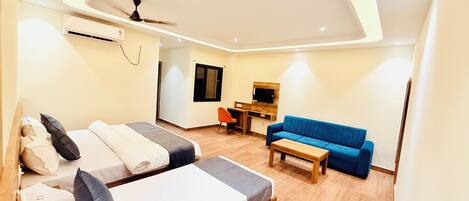A modern hotel room with two beds, a blue sofa, a wooden coffee table, a desk with a flat-screen TV, and a ceiling fan.