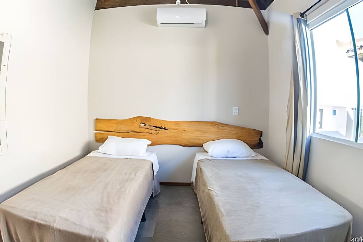 Une chambre avec deux lits simples, une tête de lit en bois, une fenêtre avec des rideaux et un climatiseur fixé au mur.