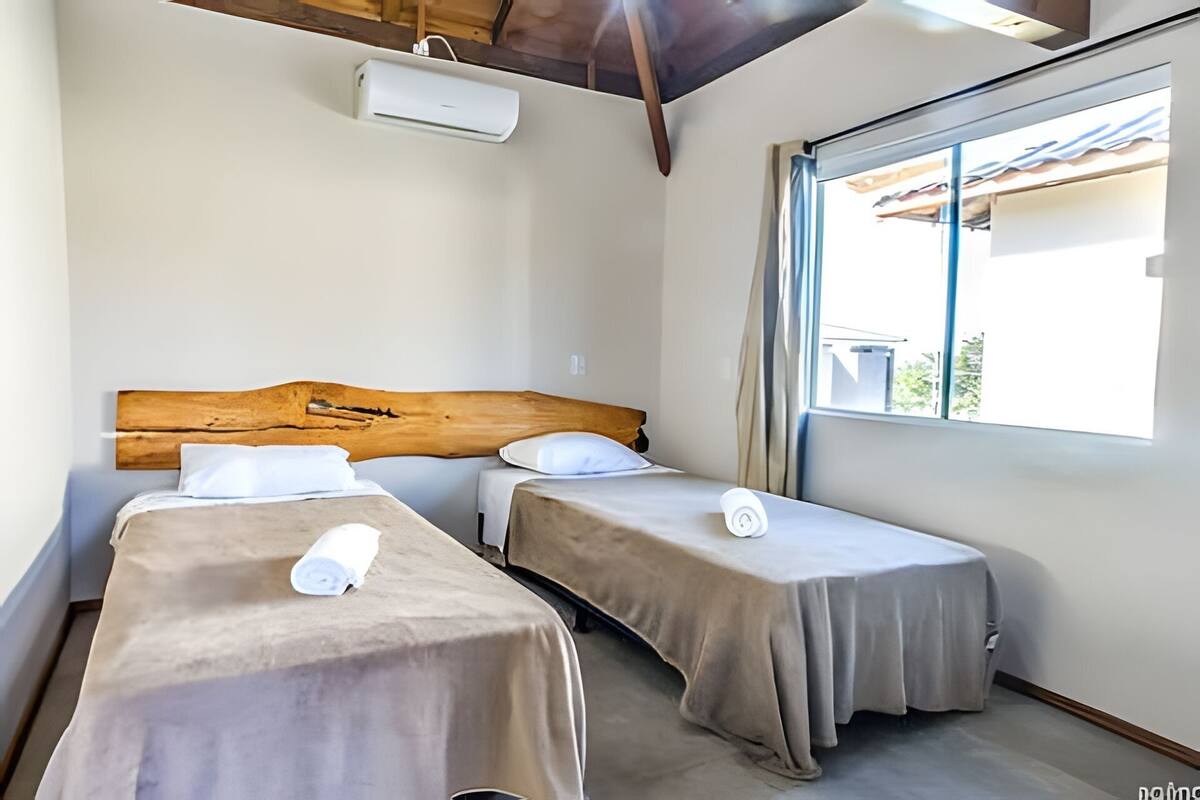 Une chambre avec deux lits simples, une tête de lit en bois, une fenêtre avec des rideaux et un ventilateur de plafond.