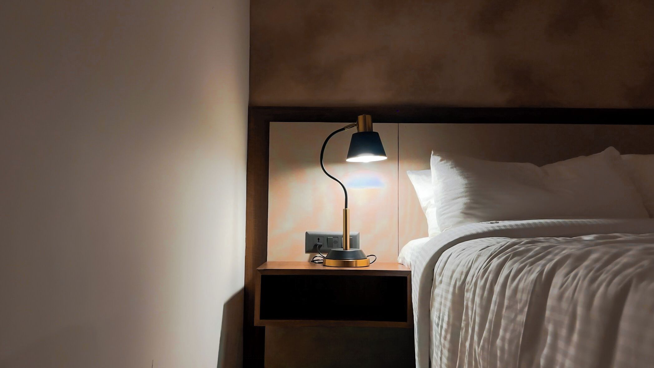 A neatly made bed with white bedding, a bedside table with a lamp, and a power outlet.
