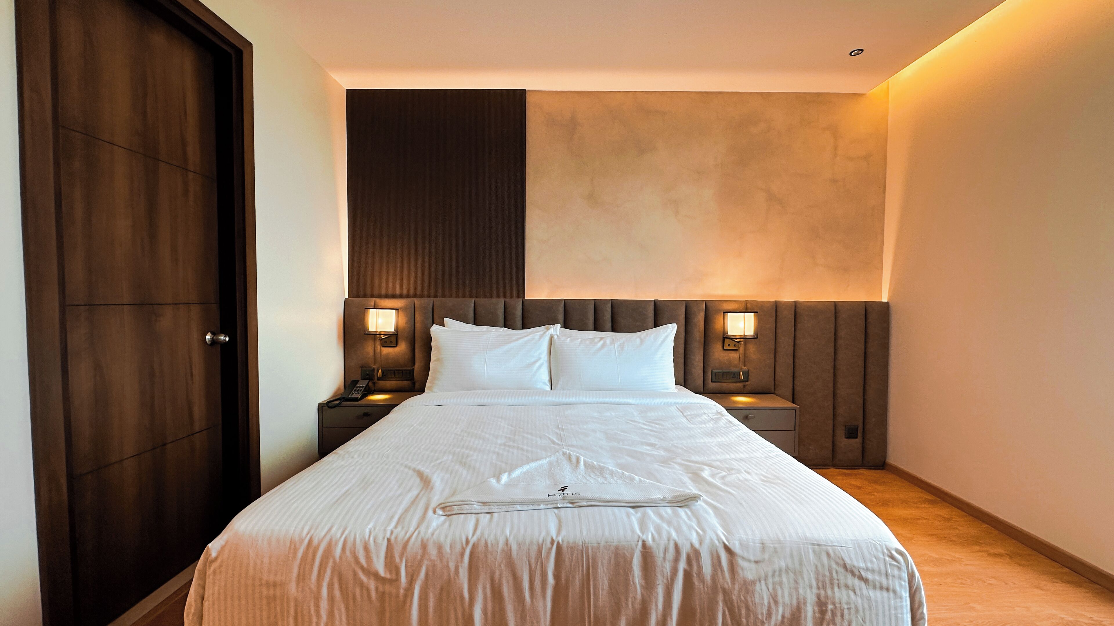 A hotel room with a large bed, two bedside tables, a wooden wardrobe, and a textured wall.