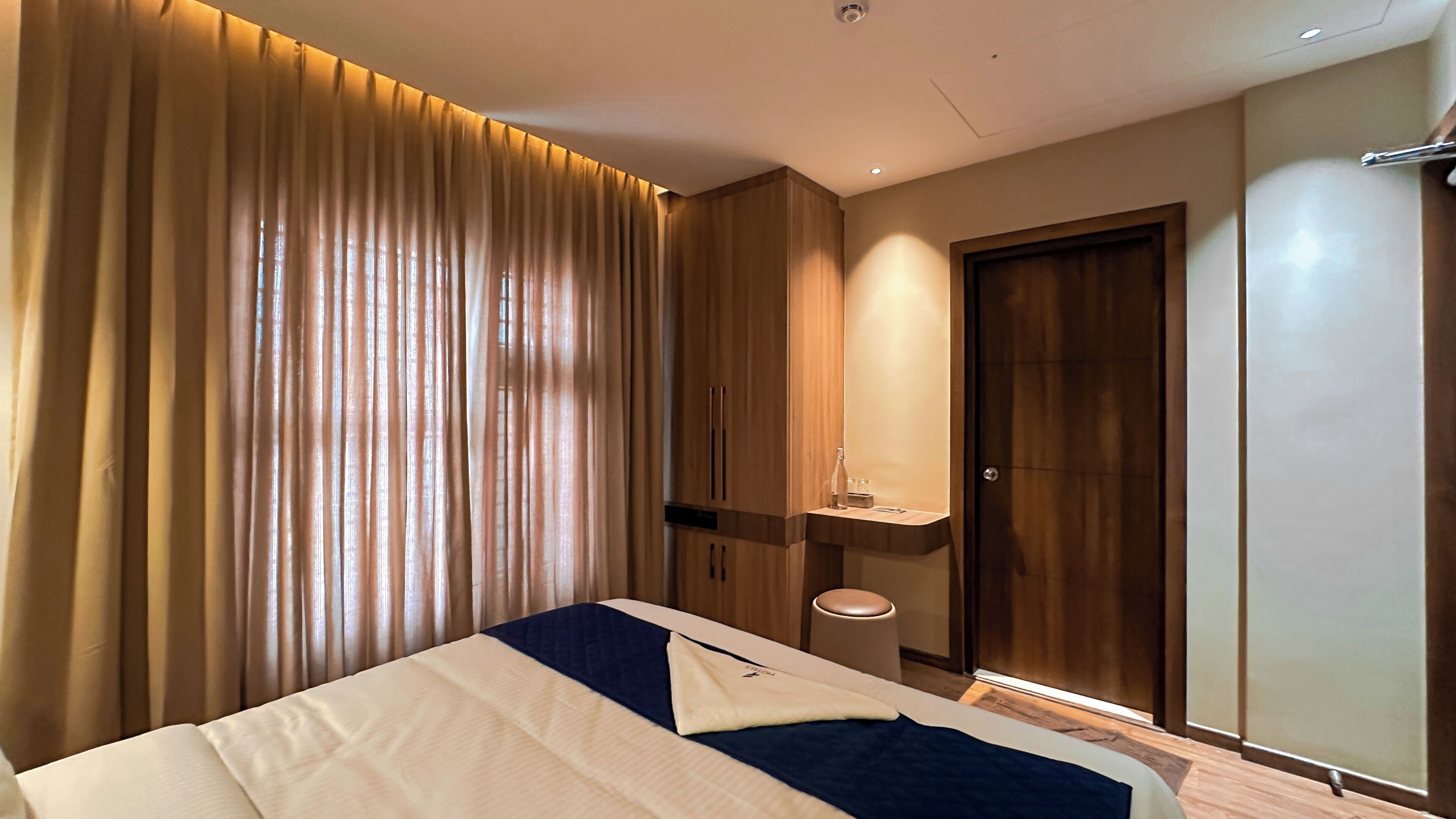 A modern hotel room with a large bed, a wooden wardrobe, a small bedside table, and a door with a mirror.