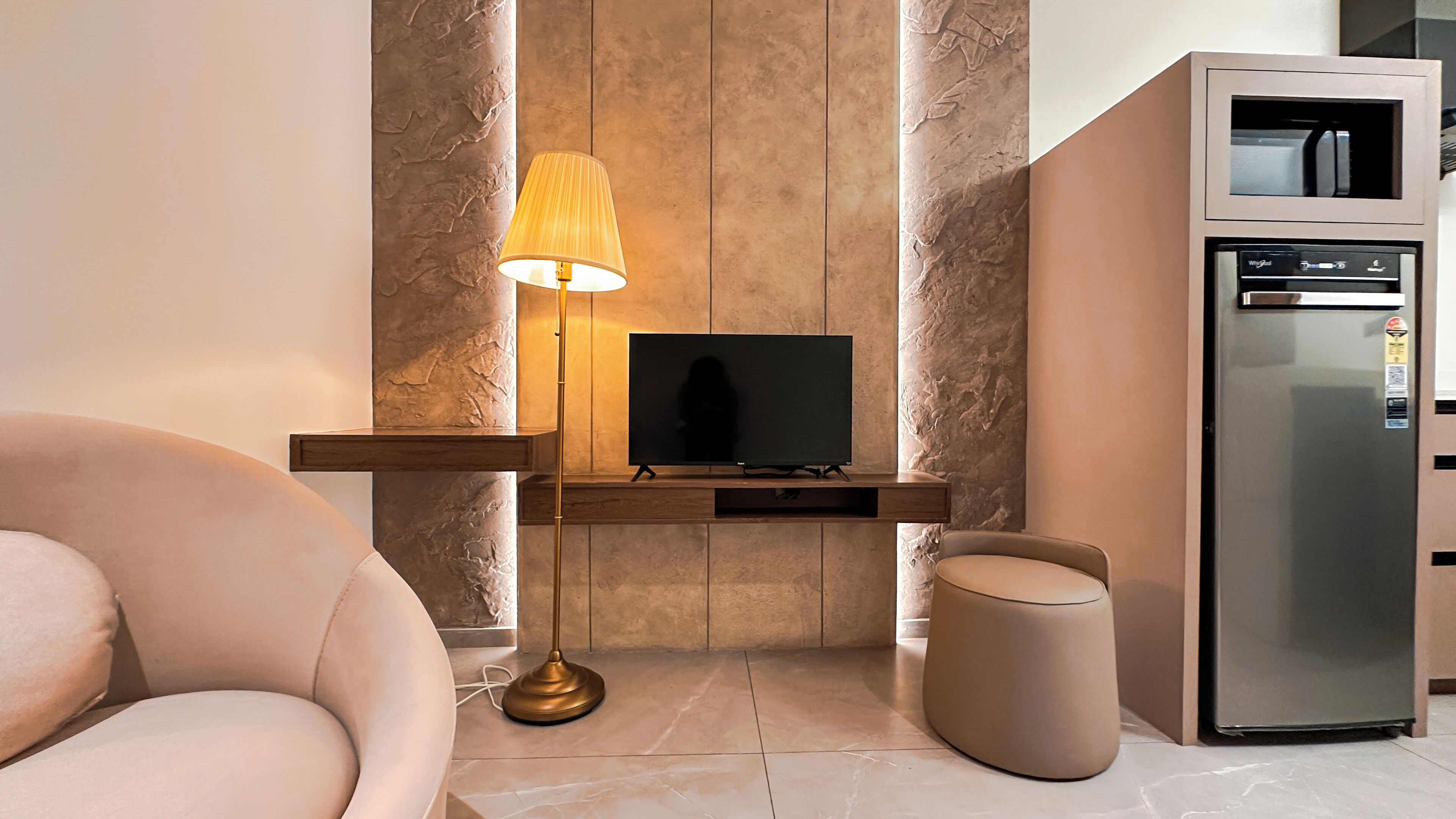 A modern living room with a flat-screen TV on a wooden console, a floor lamp, a sofa, and a built-in refrigerator.