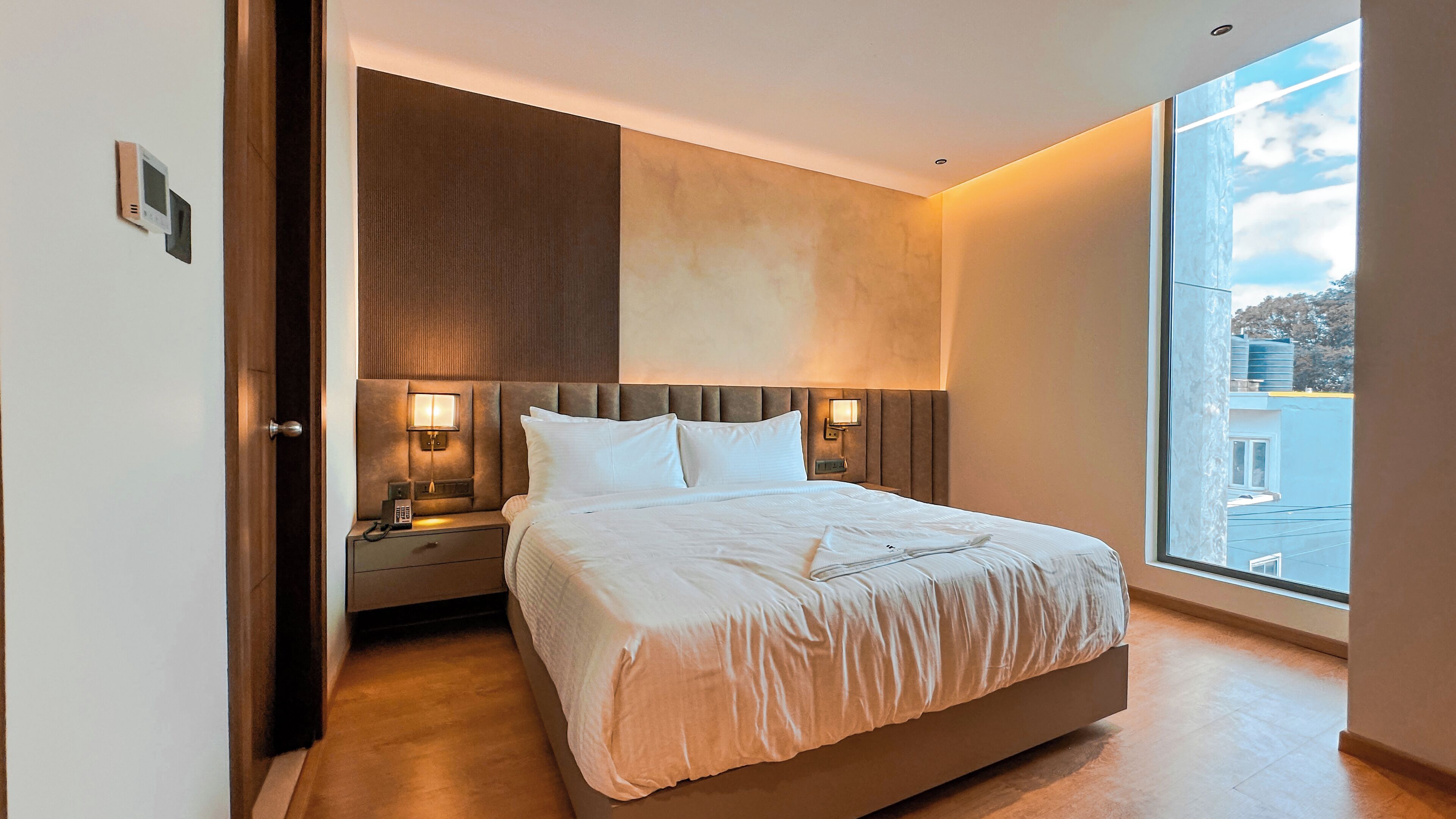Habitación de hotel moderna con una cama grande, lámparas de noche, un teléfono y vista a la ciudad a través de un ventanal amplio.