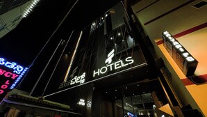 Un hôtel reconnaissable à son grand panneau « HÔTELS », à sa façade moderne en verre et à un panneau « OUVERT MAINTENANT » indiquant qu’il est possible d’y réserver une chambre.