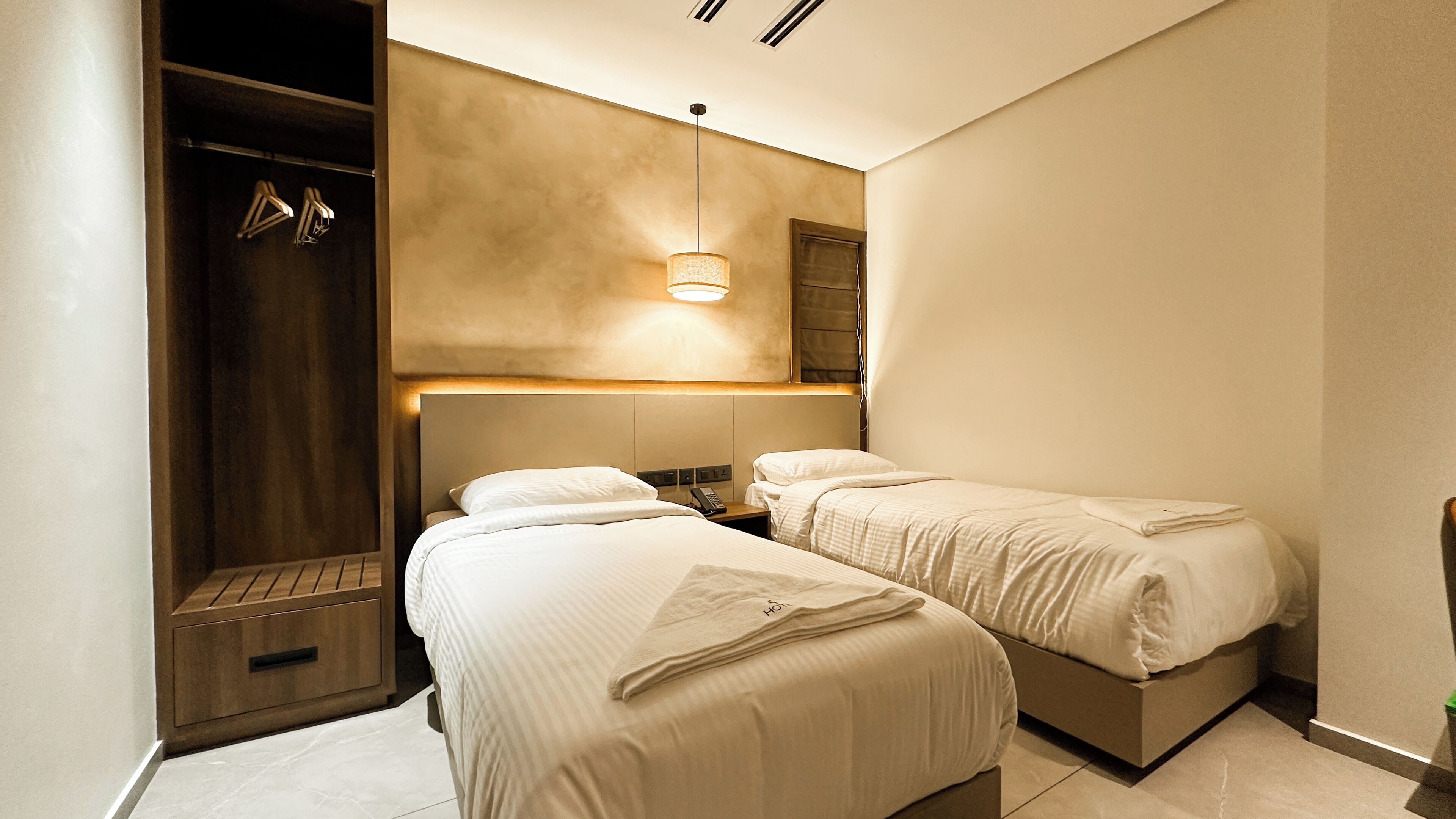 A hotel room with two beds, a wooden wardrobe, a mirror, and a wall-mounted light fixture.