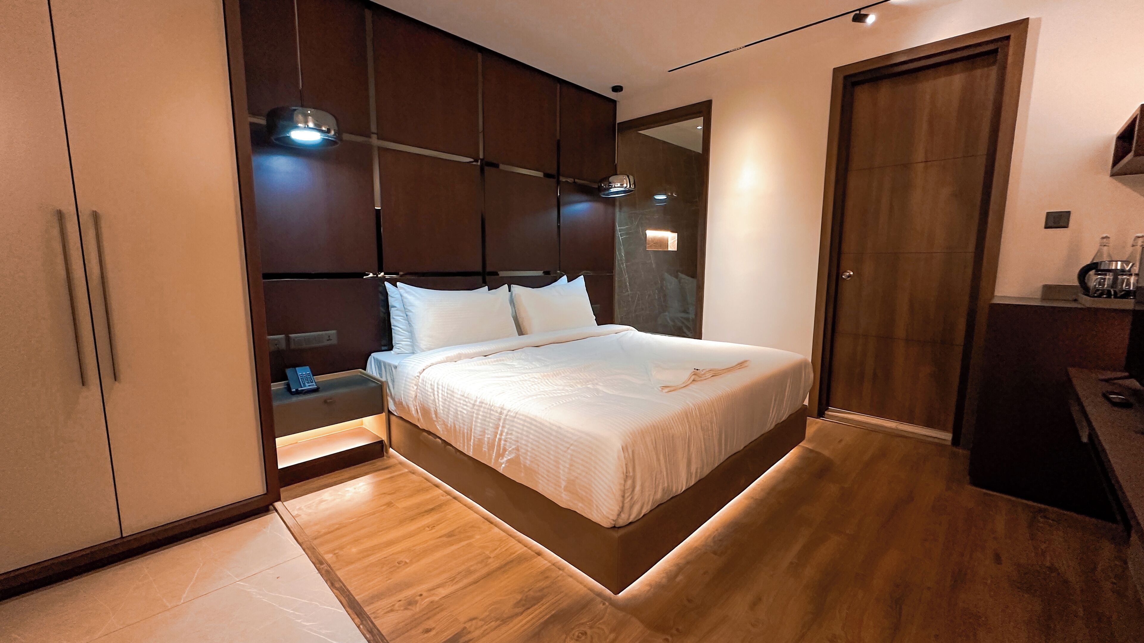 A modern bedroom with a large bed, wooden flooring, a built-in wardrobe, and a bedside table with a lamp.