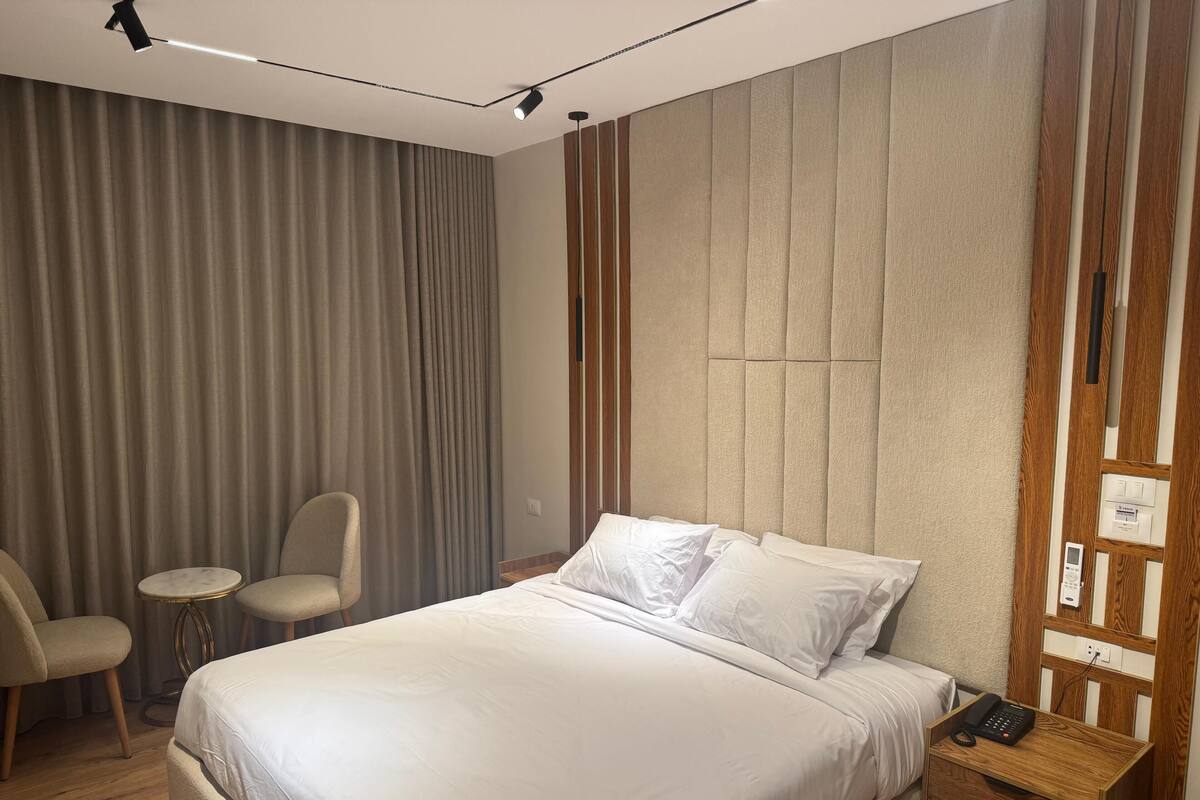 A modern hotel room with a large bed, two chairs, a small round table, and a wooden nightstand with a phone.