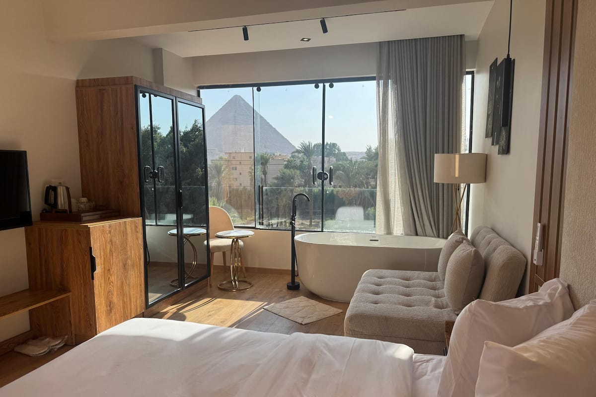 A hotel room with a large bed, a seating area with a view of a pyramid, a bathtub, and a wooden cabinet.