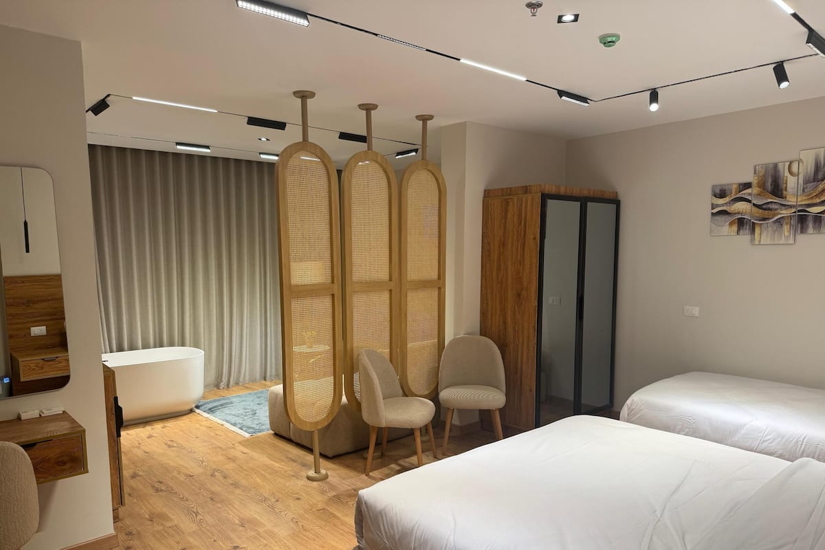 A hotel room with two beds, a sitting area with a chair, a wooden floor, a large window with curtains, and a wall-mounted artwork.