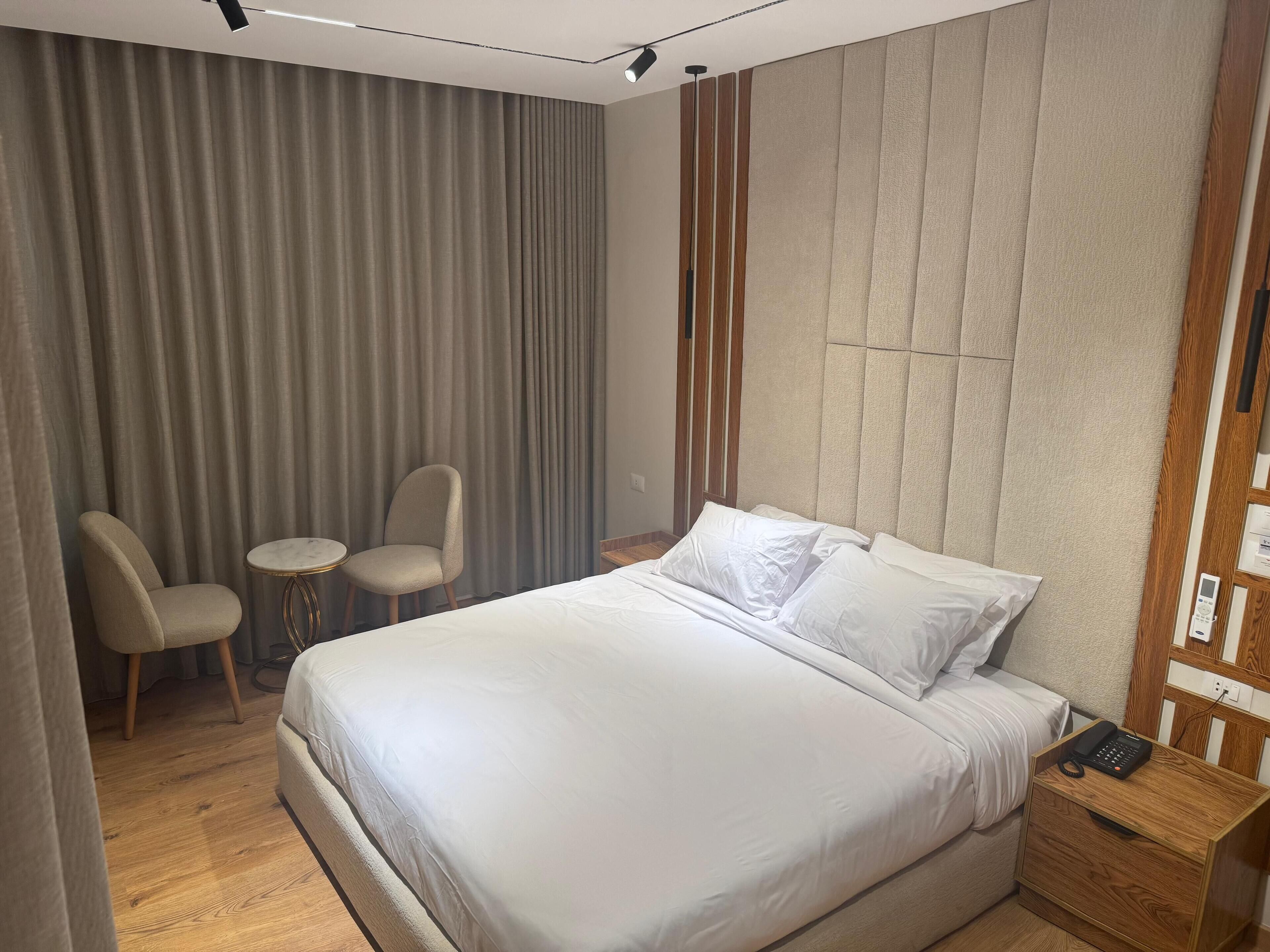 Een hotelkamer met een groot bed, twee stoelen, een klein rond tafeltje en een houten nachtkastje met een telefoon.