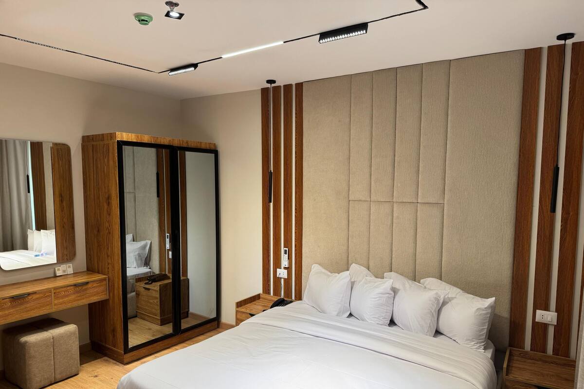 A modern hotel room with a large bed, a wooden wardrobe, a vanity with a mirror, and a bedside table.