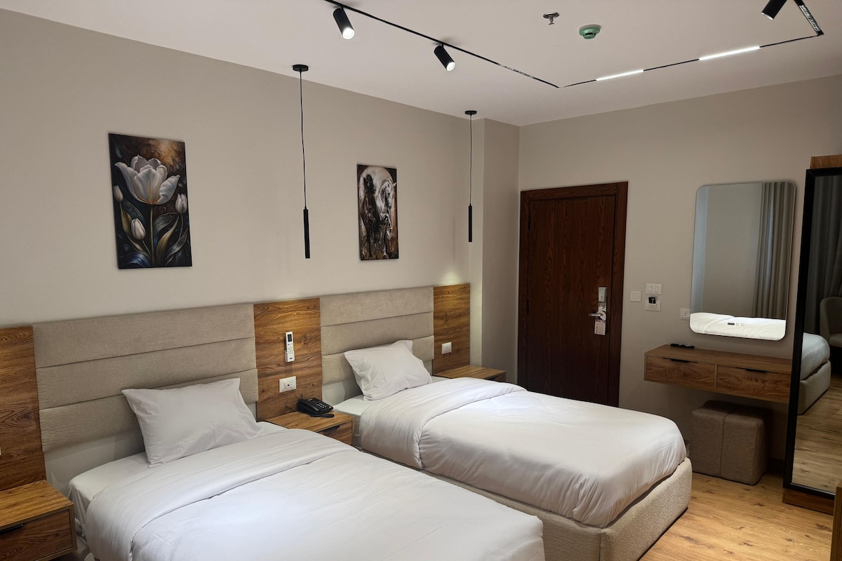 Una habitación de hotel moderna con dos camas, un cabecero de madera, una mesita de noche con teléfono, una mesita auxiliar pequeña, un espejo y suelo de madera.