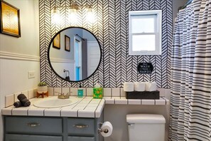 Une salle de bain avec un mur orné d’un motif en chevrons, un miroir rond, un lavabo avec deux robinets, des toilettes et un rideau de douche à rayures.
