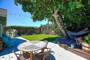 Un jardin avec une terrasse en bois, une table ronde entourée de chaises, un hamac et une petite maison de jeu.