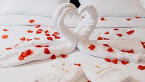 Asciugamani bianchi piegati con ricami dorati e petali di rosa rossa sparsi su un letto bianco.