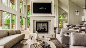 A spacious living room with a fireplace, large windows, and a television mounted above it. - Luxe Home - Heated Pool, Hot Tub & Mountain Views! (Sevier County)