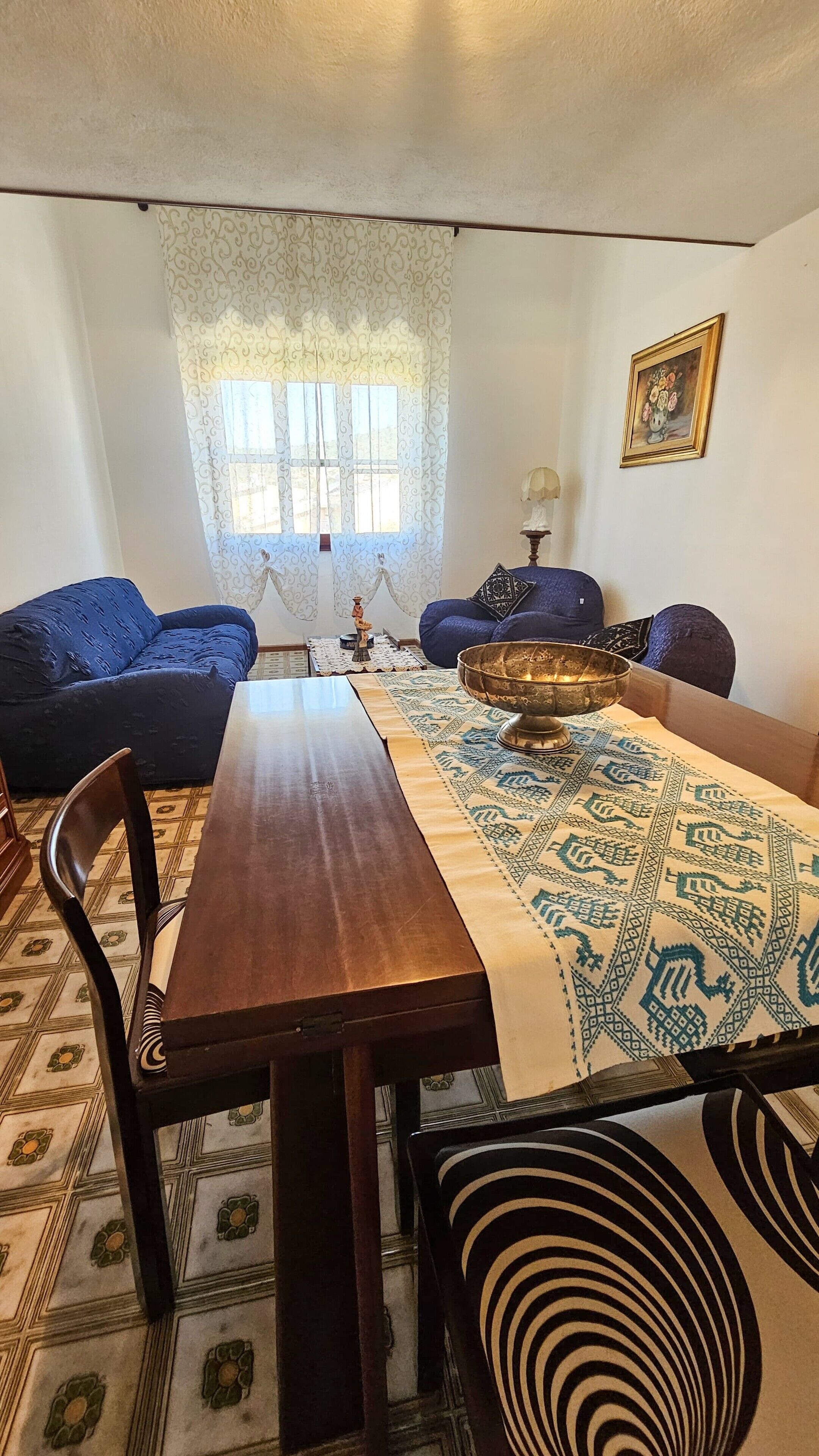 Uma área de refeições com mesa de madeira, sofás azuis, toalha de mesa estampada e uma tigela decorativa.
